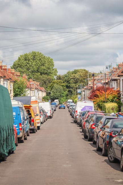 A residential street in Bowes Park shown during the daytime with overcast skies, featuring parked cars lining both sides of the narrow tarmac road. Various vehicles, including small delivery vans and private cars, are parked close to the pavement edge. In the foreground, a green cover is draped over a large item being prepared for loading, possibly furniture or boxes used for home relocation. The street is bordered by terraced houses with brick facades, pitched roofs, and small front gardens with trees and shrubs, some displaying autumn foliage. In the background, a large leafy tree dominates the scene, and overhead power lines stretch above the street. This setting illustrates typical logistical considerations for local removals, with vehicles and packing materials positioned for furniture transport and home packing during a move, as managed by companies like Man with Van Bowes Park.
