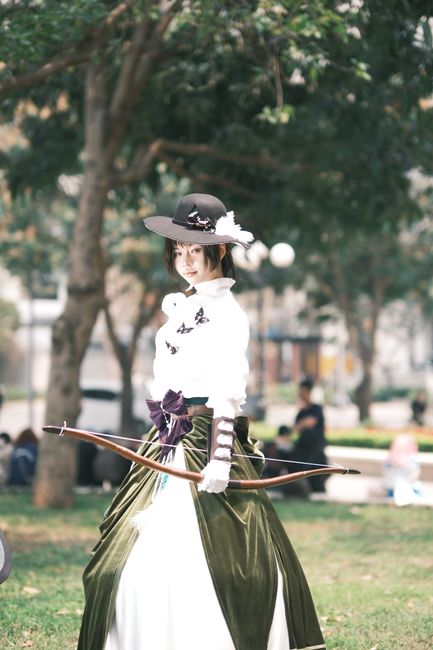 This image depicts a woman dressed in a historical or fantasy costume standing outdoors in a park setting. She is wearing a wide-brimmed black hat decorated with a white feather and flowers, and a white blouse with long, striped sleeves and butterfly embellishments. The outfit includes a dark green velvet skirt with a high waist, accented by a large purple bow, and layered with a white underskirt. She holds a bow in her left hand, with the string relaxed, and her gaze is directed towards the camera. The background features blurred trees with green foliage, park benches, and a few people in casual clothing sitting or walking on the grass. The lighting is natural, suggesting daytime with a clear, bright atmosphere. This visual could be associated with themed events or costumes, suitable for portraying character dressing in promotional images for home relocation or event planning services.