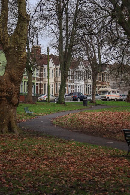 A residential street view taken from inside a park area during daytime, showing a paved walkway curving through a grassy space scattered with fallen leaves. Mature trees with bare branches border the