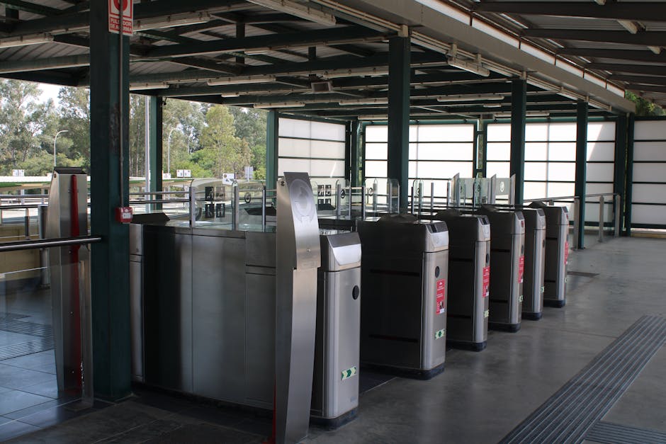 Inside Bowes Park station car park, a row of modern stainless steel ticket barriers with black rubber bases are positioned near the entrance, with clear glass panels extending upwards from the top of each barrier. To the left, a red emergency fire alarm is mounted on a dark green metal pillar. The station area features a partially covered, open-air structure with a corrugated metal roof supported by green metal beams. Behind the barriers, large tinted windows and light-colored opaque panels allow natural daylight to illuminate the space, creating a bright environment suitable for passengers or station staff. Outside, visible trees and a road provide a view into the surrounding area of Bowes Park. This station setting, captured during daytime, highlights the accessibility features and infrastructural elements typical for a UK railway station, aligned with the context of house removals and furniture transport, as part of the progressing home relocation process.