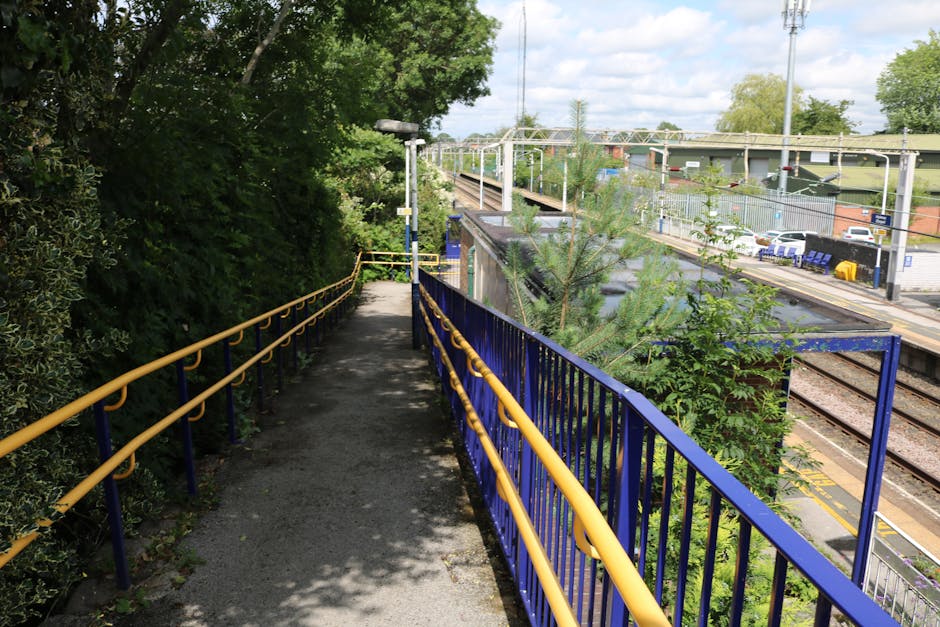 A narrow outdoor walkway beside Bowes Park railway station with a black asphalt surface and blue metal railings with yellow handrails on both sides. The pathway is shaded by leafy green trees on the left, casting dappled sunlight onto the ground. At the end of the pathway, there is a white streetlamp and a small green shrub after which the path opens to the station platform and train tracks. The station area on the right features multiple railway lines with overhead electric wiring and posts, a fenced car park with several parked vehicles, and a row of blue seating benches. The scenery includes trees, bushes, and a partly cloudy sky overhead. This setting illustrates an accessible route adjacent to the station, relevant to services offered by Man with Van Bowes Park, including home relocation and furniture transport, highlighting logistical considerations for removals and packing during moving projects.