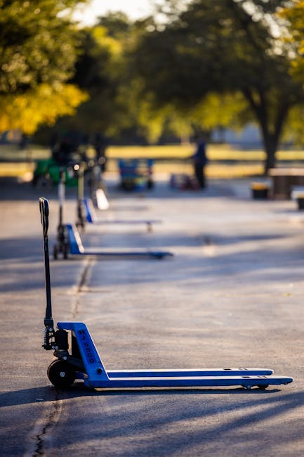 A row of blue two-wheeled hand trucks or dollies lined up on an empty outdoor parking area or street, with a smooth asphalt surface marked by faint white lines. The first dolly is positioned close to the camera, with its platform flat on the ground and the handle upright, designed for lifting and transporting heavy or bulky items such as furniture during home relocation or furniture transport. In the background, there are blurred trees with green and yellow foliage, and some indistinct figures and objects further along the street, suggesting a park or residential environment. The lighting indicates daytime with natural sunlight casting minimal shadows, and the scene reflects preparations related to packing and moving services provided by Man with Van Bowes Park, highlighting the logistics involved in loading large items onto a vehicle for house removals.