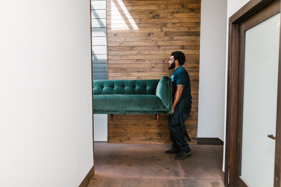 A man wearing a dark teal uniform is engaged in a home relocation process inside a room with white walls and a wooden panel accent wall. He is lifting a large, upholstered green velvet sofa, carefully carrying it through a doorway onto a carpeted area. The sofa appears to be well-padded with tufted design, and the man is holding it securely, ensuring proper handling during the furniture transport. To the right of the image, a partially visible glass door or window frame is present, providing natural light that illuminates the scene. Nearby, a white side table or small storage unit is seen beneath the sofa. The environment looks organized, with a focus on careful packing and handling, supported by the use of proper lifting techniques and furniture blankets, indicative of professional removals services from Man with Van Bowes Park. This process reflects a typical part of furniture transport and packing during house removals designed to protect items and facilitate efficient household moving.