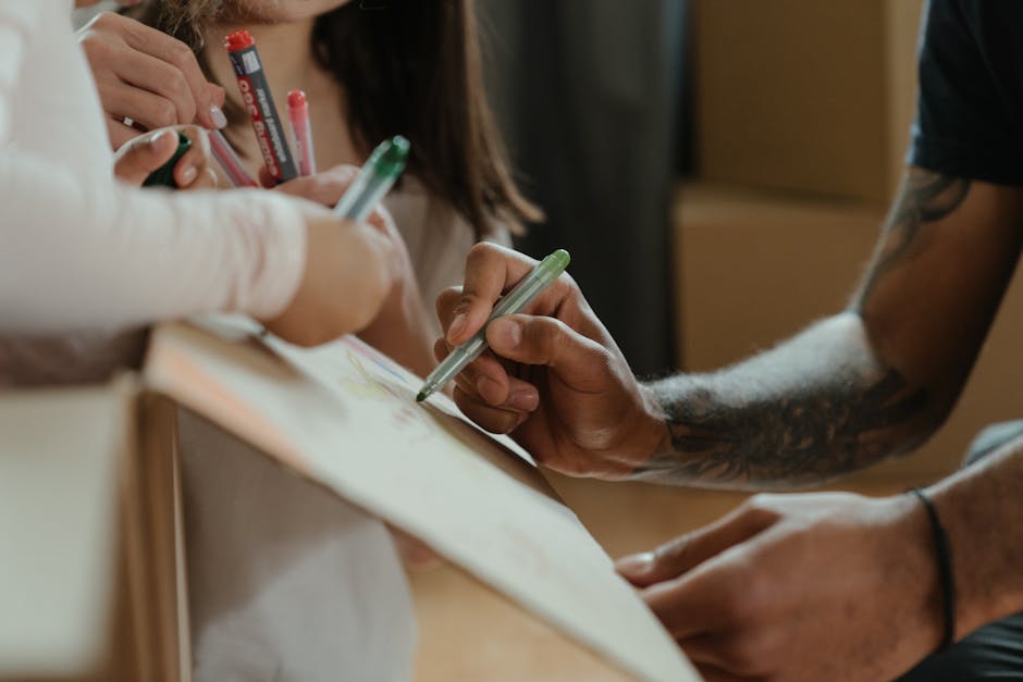 A person with a tattooed arm is seen using a green marker to label cardboard boxes in preparation for a home relocation, with children and adults involved in packing activities visible in the backgrou