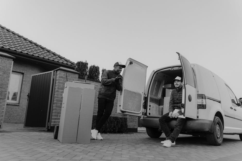 Two movers from Man with Van Bowes Park are engaged in a home relocation process outside a residential property during daytime. One mover, standing next to an open van, is holding a clipboard or devic