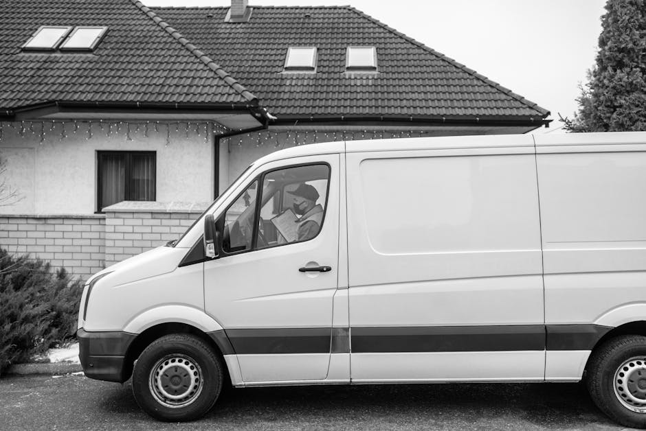 Two movers from Man with Van Bowes Park are engaged in a home relocation process outside a residential property during daytime. One mover, standing next to an open van, is holding a clipboard or devic