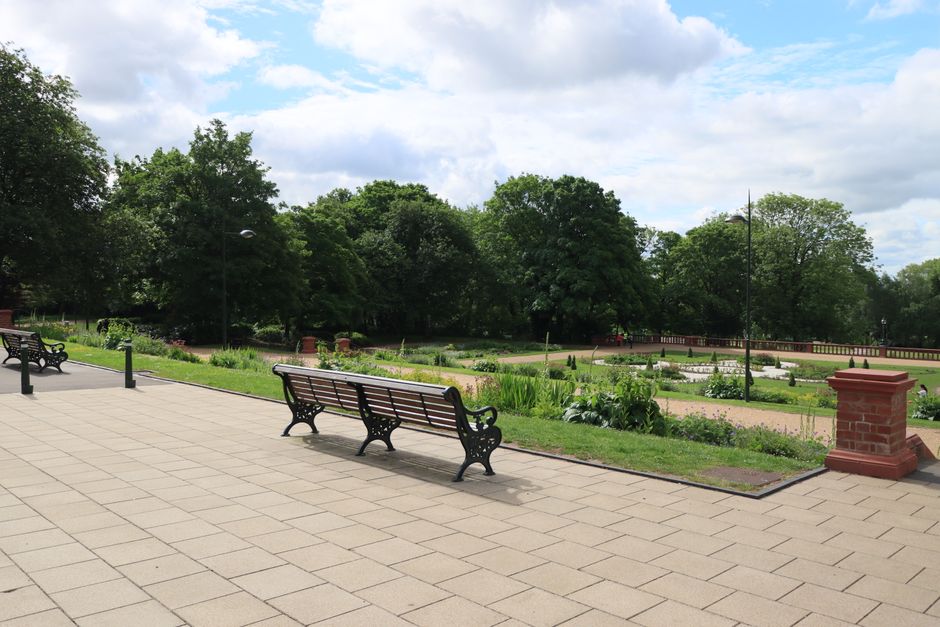 A park scene viewed through tree branches, featuring a rectangular pond with a few ducks swimming on the water's surface, surrounded by a paved walkway. In the background, there are several people walking and sitting on benches, some engaged in conversations or enjoying the scenery. Behind the park, there are trees with blooming or leafless branches, a small hill with a black metal fence, and residential houses partially visible beyond the foliage. The scene is illuminated by natural daylight, with clear weather and a peaceful, urban park atmosphere. This image relates to the residential area where house removals or relocation services, such as those provided by Man with Van Bowes Park, may be relevant during home relocation or moving processes involving outdoor surroundings.