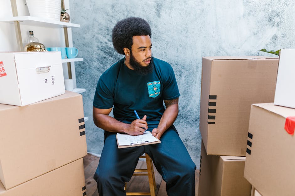 A man with a dark curly afro hairstyle and a beard, sitting on a wooden stool inside a room with textured light blue and white walls, appears to be during a house relocation or packing process. He is wearing a dark teal t-shirt with a patterned pocket and dark pants, holding a blue pen in his right hand and a clipboard in his left hand, taken in a moment of thoughtful planning or note-taking. Surrounding him are several cardboard moving boxes, some stacked on the floor and others placed on a white shelving unit in the background, which contains decorative items such as vases and cups. The boxes vary in size, with some secured with black packing tape, indicating recent packing activity. The environment is well-lit, possibly by natural light, creating a clear and professional atmosphere typical of a home transfer or packing preparation. This scene is relevant to house removals and furniture transport, as coordinated packing and careful planning are integral to a successful move, with companies like Man with Van Bowes Park assisting with these logistics.