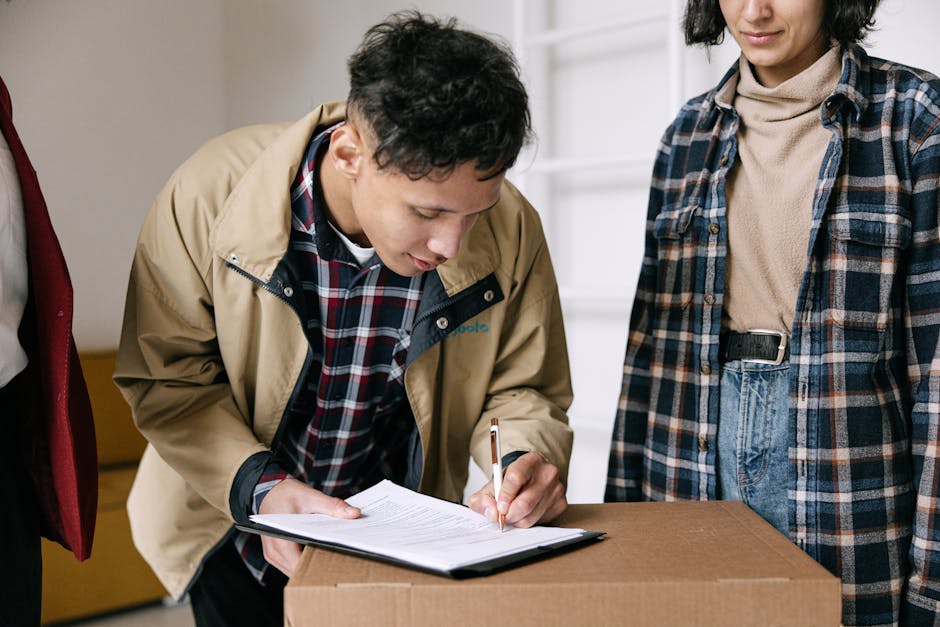 A man with a dark curly afro hairstyle and a beard, sitting on a wooden stool inside a room with textured light blue and white walls, appears to be during a house relocation or packing process. He is wearing a dark teal t-shirt with a patterned pocket and dark pants, holding a blue pen in his right hand and a clipboard in his left hand, taken in a moment of thoughtful planning or note-taking. Surrounding him are several cardboard moving boxes, some stacked on the floor and others placed on a white shelving unit in the background, which contains decorative items such as vases and cups. The boxes vary in size, with some secured with black packing tape, indicating recent packing activity. The environment is well-lit, possibly by natural light, creating a clear and professional atmosphere typical of a home transfer or packing preparation. This scene is relevant to house removals and furniture transport, as coordinated packing and careful planning are integral to a successful move, with companies like Man with Van Bowes Park assisting with these logistics.