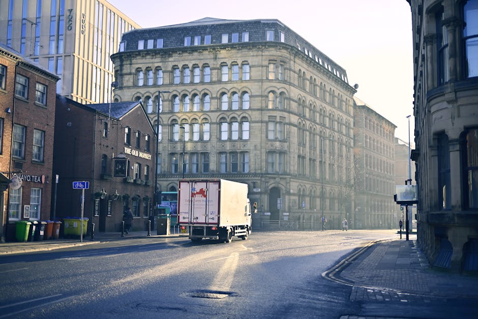 A small white moving truck with a closed cargo area is parked on a wide city street during daylight hours, positioned near a stone building with multiple large windows and ornate architectural features. To the left, there are brick buildings, one with a sign reading 'The Old Monastery,' and several recycling bins of different colours near the sidewalk. The truck is aligned roughly in the middle of the scene, facing away from the camera, with the rear doors closed. The street appears calm with minimal traffic, and a few pedestrians are visible in the distance. The environment is well-lit with natural daylight, creating slight shadows on the pavement. The scene captures an urban setting typical of house removals or furniture transport activities, with the truck ready for a home relocation, providing a visual context for professional moving services such as those offered by Man with Van Bowes Park.