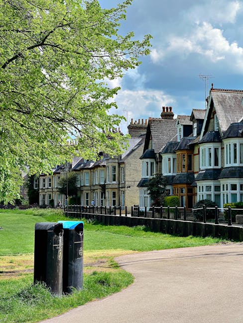 A row of traditional terraced houses with bay windows and slate roofs, situated behind a wooden fence in a residential area. In the foreground, there are two black rubbish bins on a grassy verge next to a paved footpath, partially shaded by a large leafy tree with bright green foliage. The sky is partly cloudy with patches of blue visible. The scene depicts a peaceful neighborhood environment, suitable for house removals and home relocations. The natural lighting highlights the details of the houses, the greenery, and the street infrastructure, creating a calm setting for furniture transport or packing and moving activities in Bowes Park, as managed by Man with Van Bowes Park.