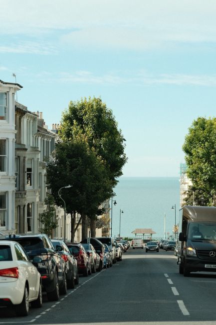 A residential street in Bowes Park, featuring a row of parked cars on both sides of the road with a variety of compact and larger vehicles, including a white car and a van visible at the right edge. Along the street, there are white terraced houses with classic Victorian-style architectural details, such as bay windows and decorative facades. Two lush, green trees line the sidewalk, providing partial shade and contrast to the urban setting. In the background, the street slopes downward toward a view of the sea, with a small pavilion or shelter structure visible near the water's edge. The sky is clear with a few wispy clouds, and the scene appears well-lit, suggesting a bright day. This setting captures the typical environment for home relocation services, with potential space for loading and unloading furniture and boxes, as handled by companies like Man with Van Bowes Park during packing and moving operations.