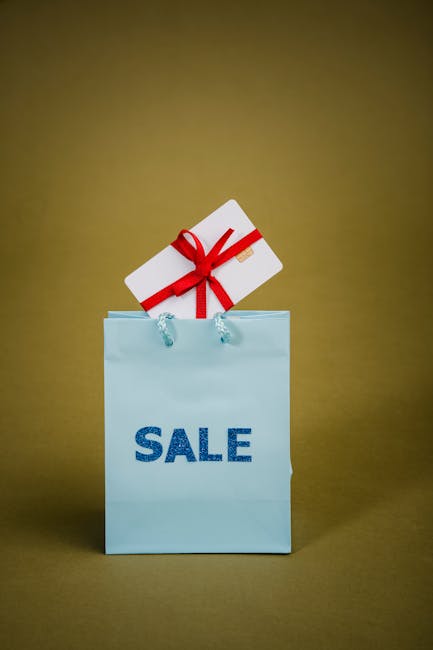 A light blue paper shopping bag with the word 'SALE' printed in dark blue letters on the front, placed against a plain, muted yellow background. The bag has two thin, white rope handles. Inside the bag, there is a rectangular white box wrapped with a bright red ribbon tied into a bow, with the ribbon extending over the edges of the box. The box is positioned at an angle, leaning slightly to the right, suggesting it has been recently placed into or is being removed from the bag. The setting is neutral and simple, emphasizing the promotional or discounted sale theme commonly associated with retail, moving, or packing contexts, which are relevant to home relocation or furniture transport scenarios often managed by companies like Man with Van Bowes Park.